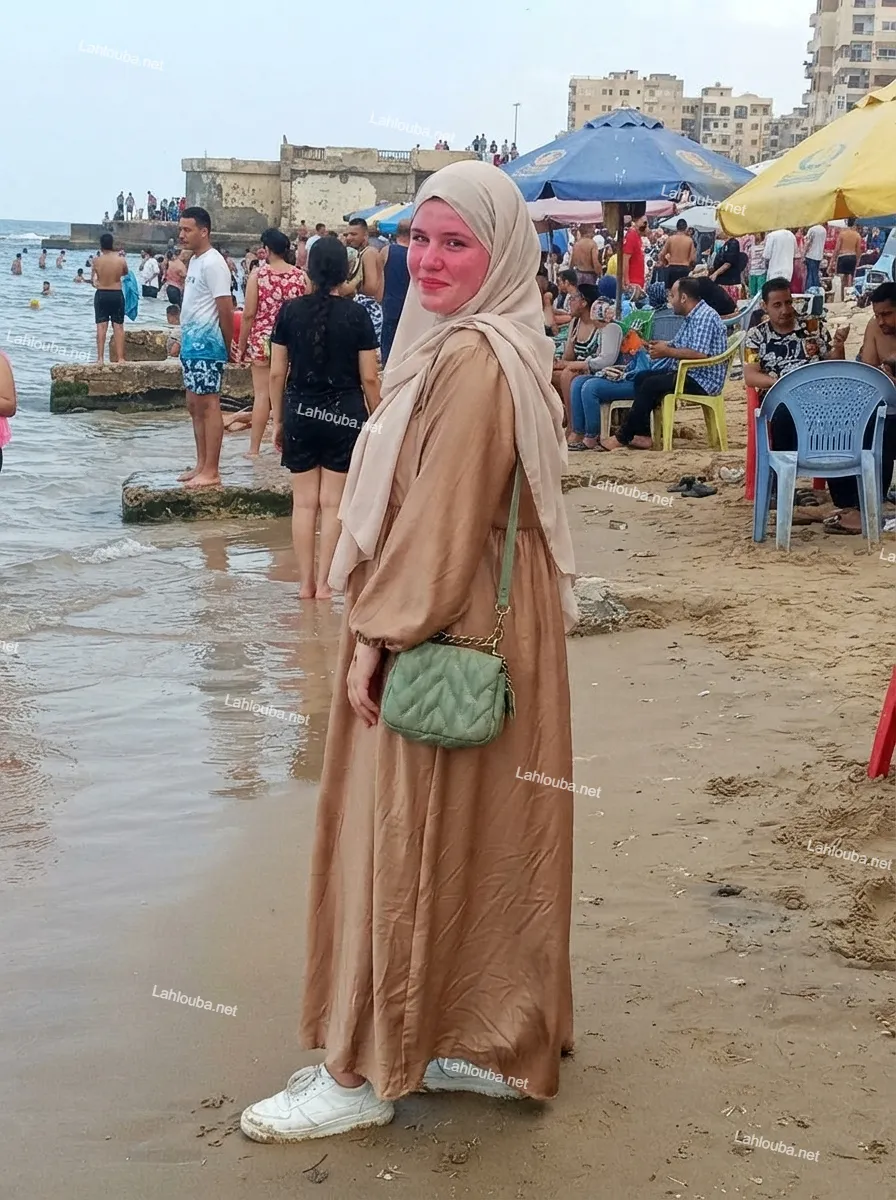 فتاة محجبة بفستان بني وحقيبة خضراء على شاطئ البحر