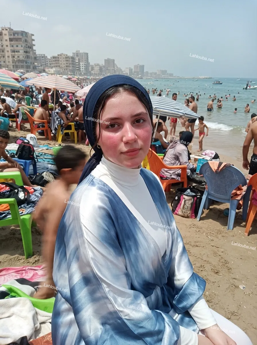 فتاة مسلمة ترتدي كيمونو بتصميم Tie-dye أزرق وأبيض وحجاب أزرق داكن على شاطئ مزدحم.