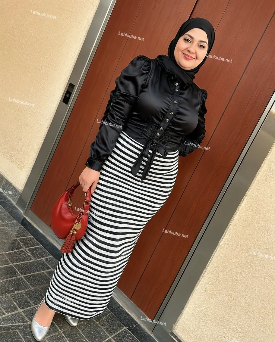 إمرأة ترتدي بلوزة ساتان سوداء بأكمام بوفان وتنورة طويلة مخططة بالأبيض والأسود مع حقيبة حمراء وحجاب أسود
