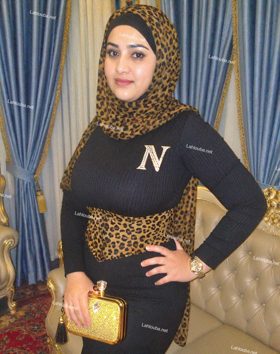 شابة ترتدي فستاناً أسود مخططاً مطرزاً بحرف N كبيرة مع غطاء رأس بنمط طباعي وحقيبة يد ذهبية لامعة