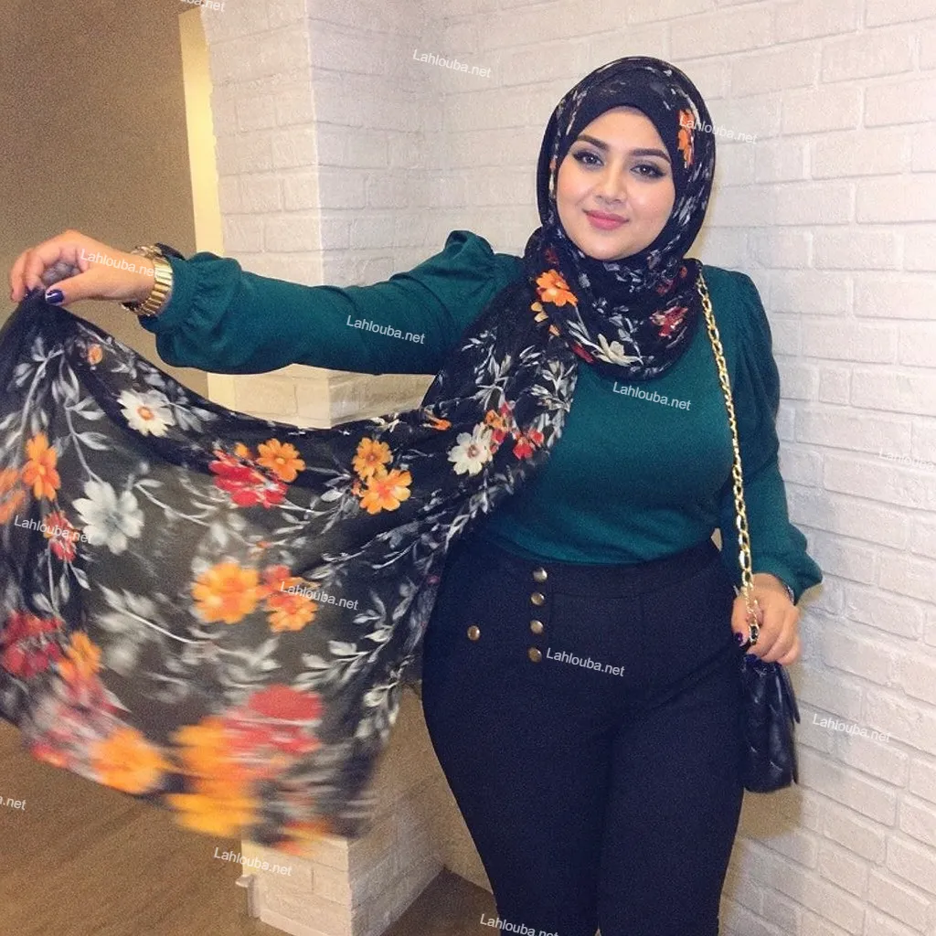 سيدة ترتدي قميصاً أخضر داكناً مع حجاب مطبوع بالورود وبنطلون أسود عالي الخصر وحقيبة بسلسلة ذهبية