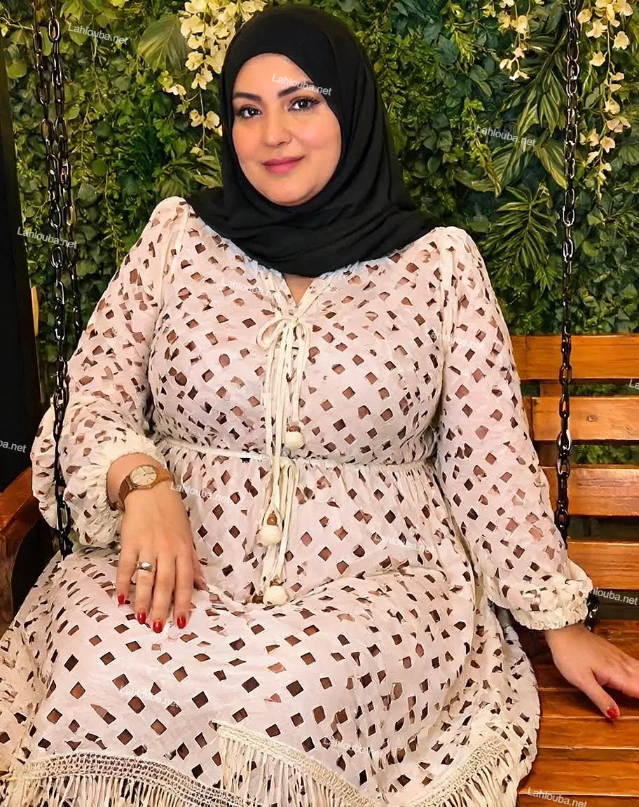 مرأة ترتدي فستان أبيض بتطريز مخرمي هندسي مع حجاب أسود على مقعد خشبي أمام نباتات خضراء
